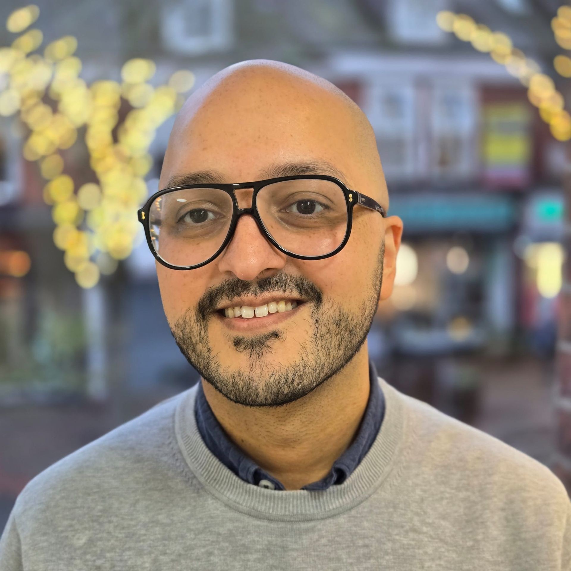 Person wearing a grey sweatshirt standing outdoors with blurred Christmas lights and buildings in the background.